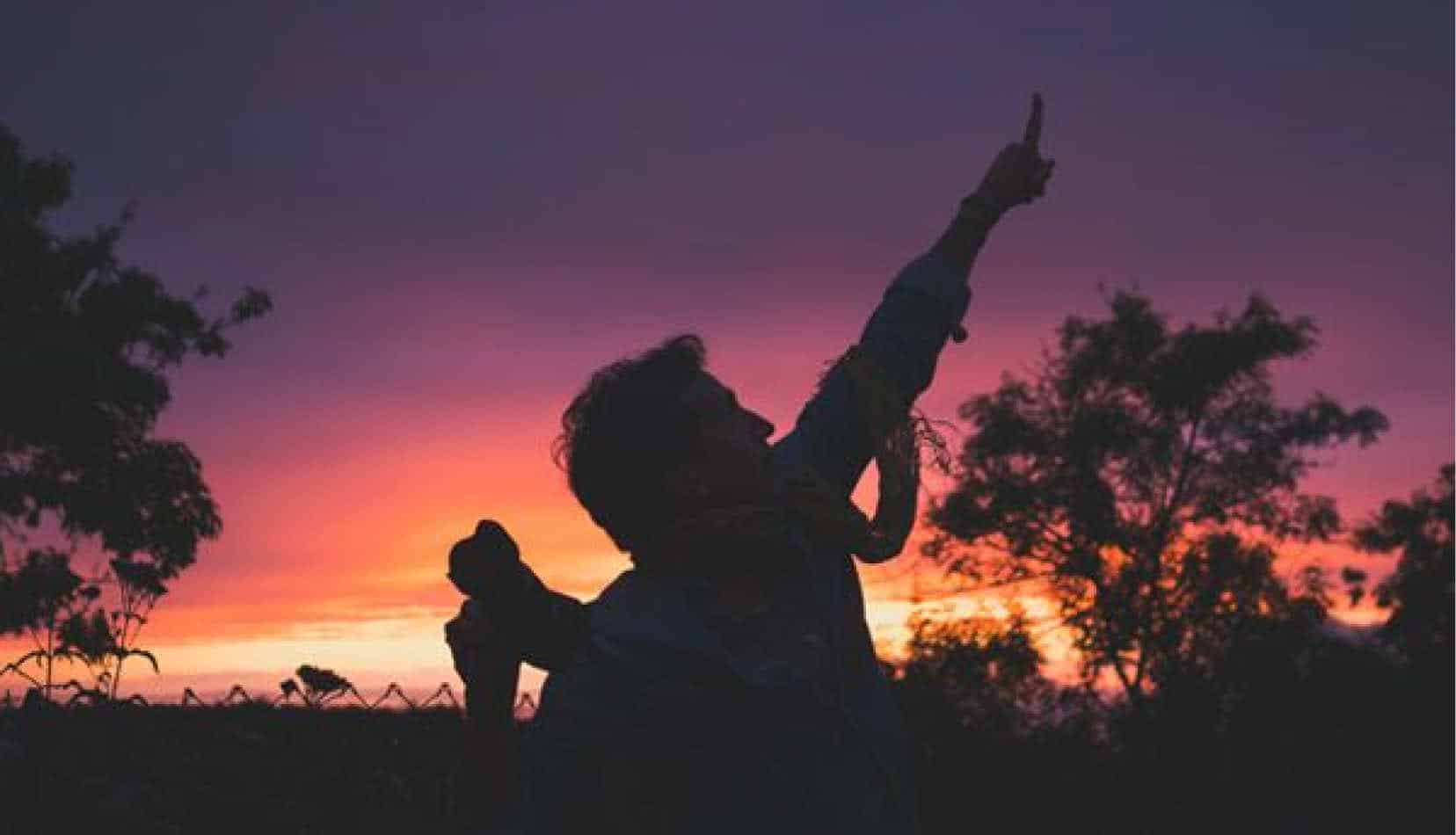 A man silhouetted by the sunset points into the sky
